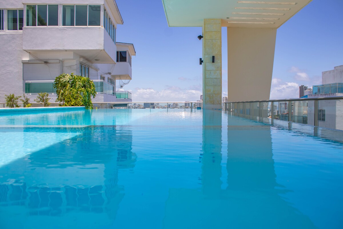 An inviting rooftop pool is presented, featuring a stunning view of the sky and cityscape. The pool's clear, tranquil waters create a serene environment, framed by modern architectural elements and green landscaping.