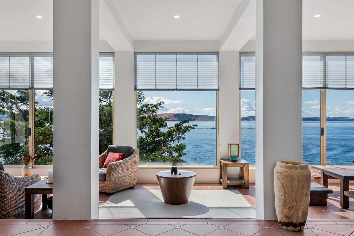 A bright living area features large windows that frame views of the water and landscape. Comfortable seating is arranged around a central wooden coffee table. Natural light enhances the spacious feel, while the surrounding greenery adds a touch of nature.