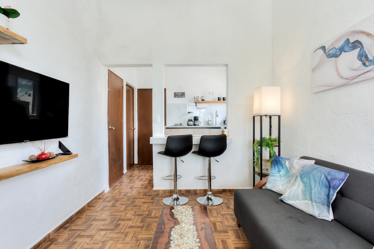 A living area showcases a modern sofa with decorative pillows, positioned near a wall-mounted television. A small bar area with two stools is visible, along with an open kitchen space in the background. Natural light illuminates the room through windows, enhancing the ambiance.
