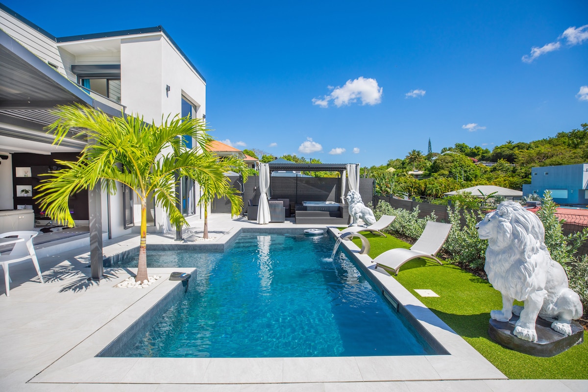 An outdoor area features a sleek rectangular pool surrounded by white lounge chairs and a tropical palm tree. Two decorative lion statues are positioned at the pool's edge, with a shaded seating area visible in the background under clear blue skies.