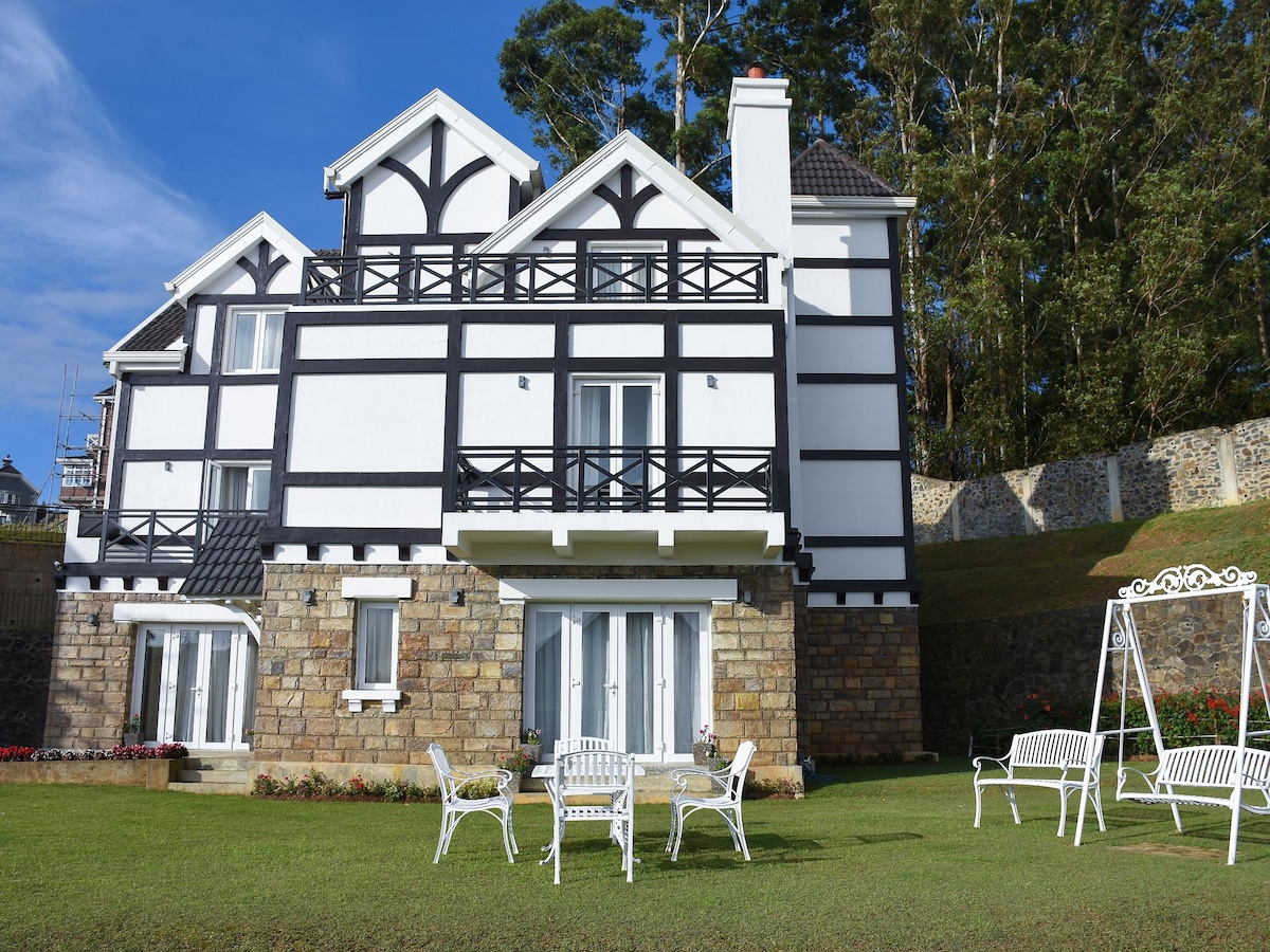 A charming three-story cottage showcases a distinctive black and white facade, framed by lush greenery. A well-maintained lawn features white outdoor furniture arranged around a small table, inviting relaxation, while large glass doors provide access to the outdoor space.