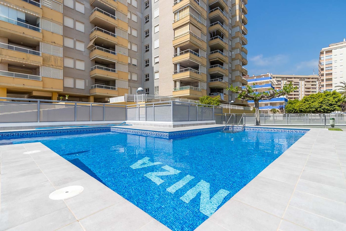 A tranquil swimming pool is surrounded by spacious concrete pathways and neatly trimmed landscaping. Clear blue water reflects the sunlight, while the nearby residential buildings provide a backdrop. The pool area is designed for relaxation and leisure, offering a welcoming environment.