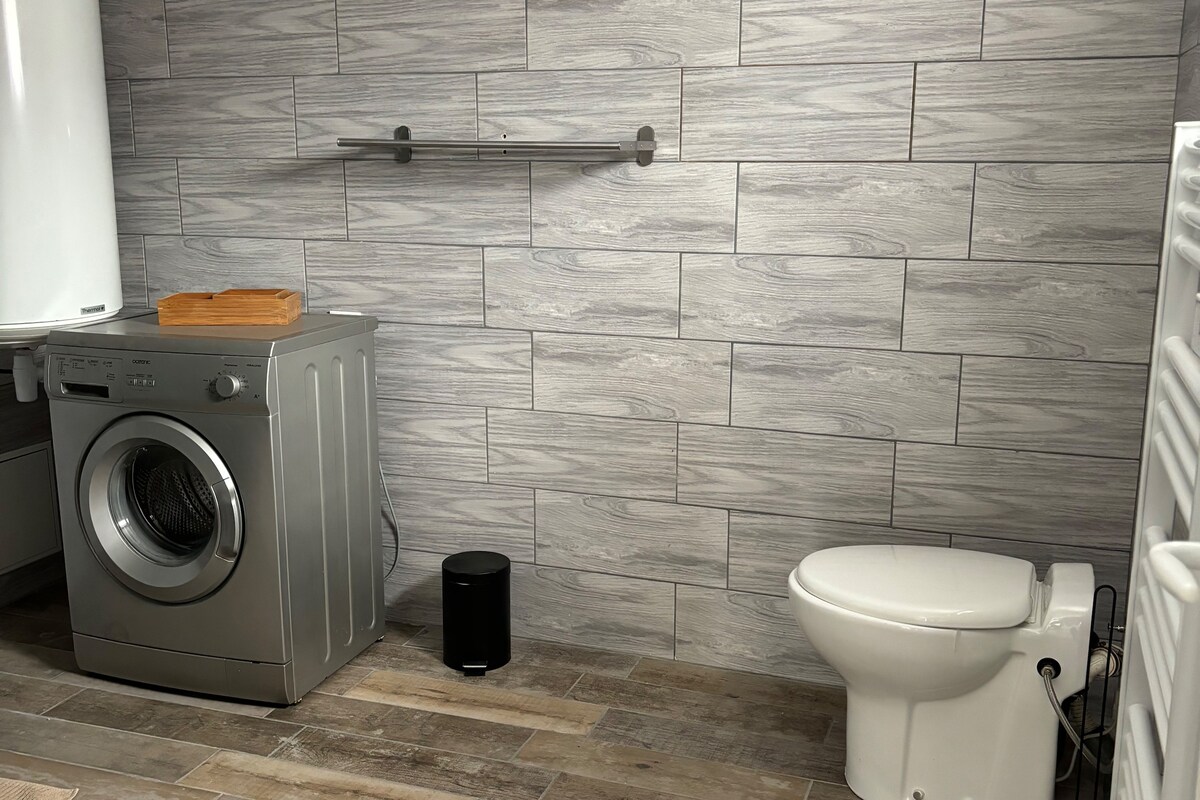A neutral-colored bathroom features modern tiles with a wood-like appearance. A washing machine is positioned beside a shelf. A sleek black trash can is located on the floor next to a white toilet, contributing to a clean and functional atmosphere.