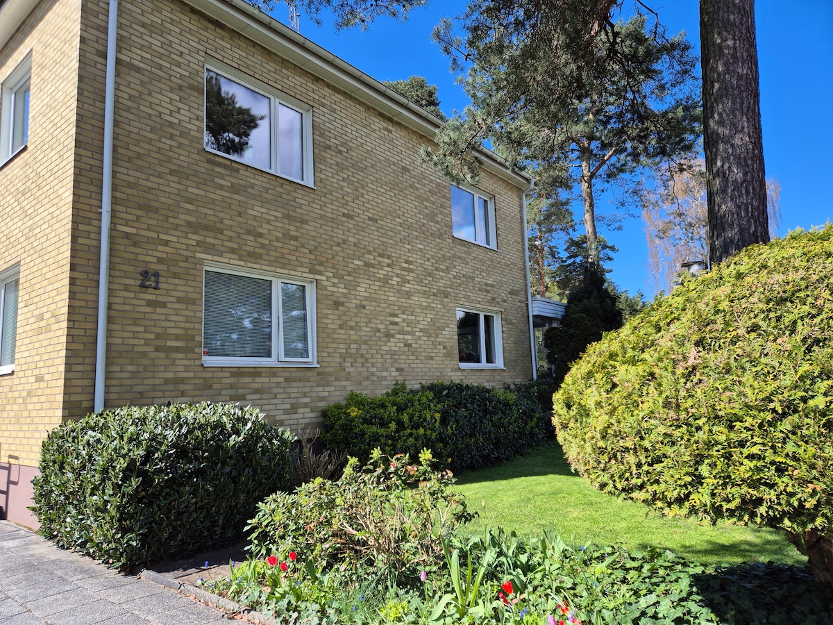 A well-maintained exterior of a two-story building is shown, featuring tan brick walls and large windows. The surrounding garden displays lush greenery, including neatly trimmed hedges and colorful flower beds. Tall trees provide shade, creating a serene outdoor environment.