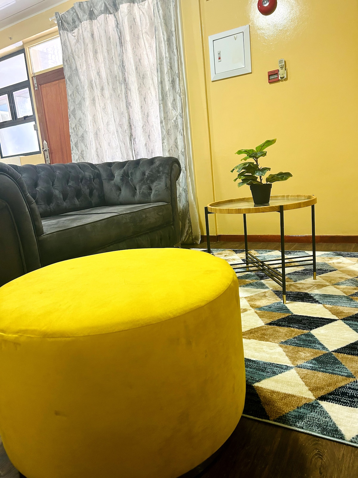 A plush grey couch is complemented by a round yellow ottoman and a small, circular coffee table featuring a potted plant. A geometric-patterned area rug adds texture to the floor, while soft curtains frame a nearby window, allowing natural light to enter the room.