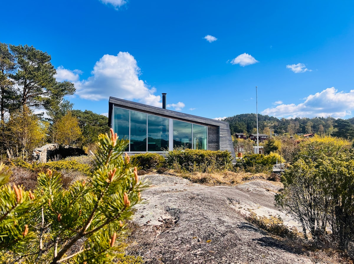 A modern architectural cabin is depicted against a clear blue sky. Large glass windows reflect the surroundings, showcasing the panoramic views. Natural vegetation frames the building, blending it harmoniously with the rocky landscape.