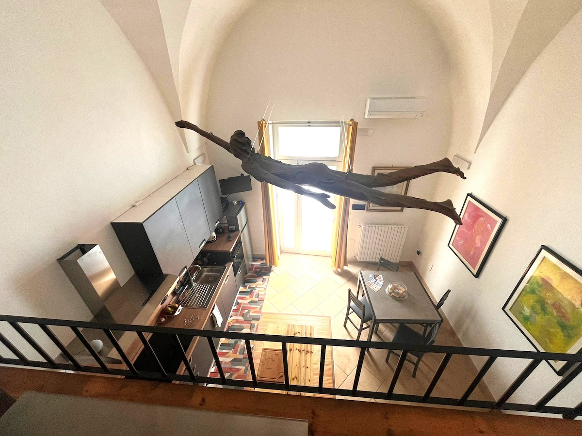 An overhead view showcases a modern kitchen with dark cabinetry and stainless steel appliances. A dining area with a table and chairs is visible, complemented by artistic wall decor. Natural light enters through large windows, enhancing the open space beneath a high ceiling.