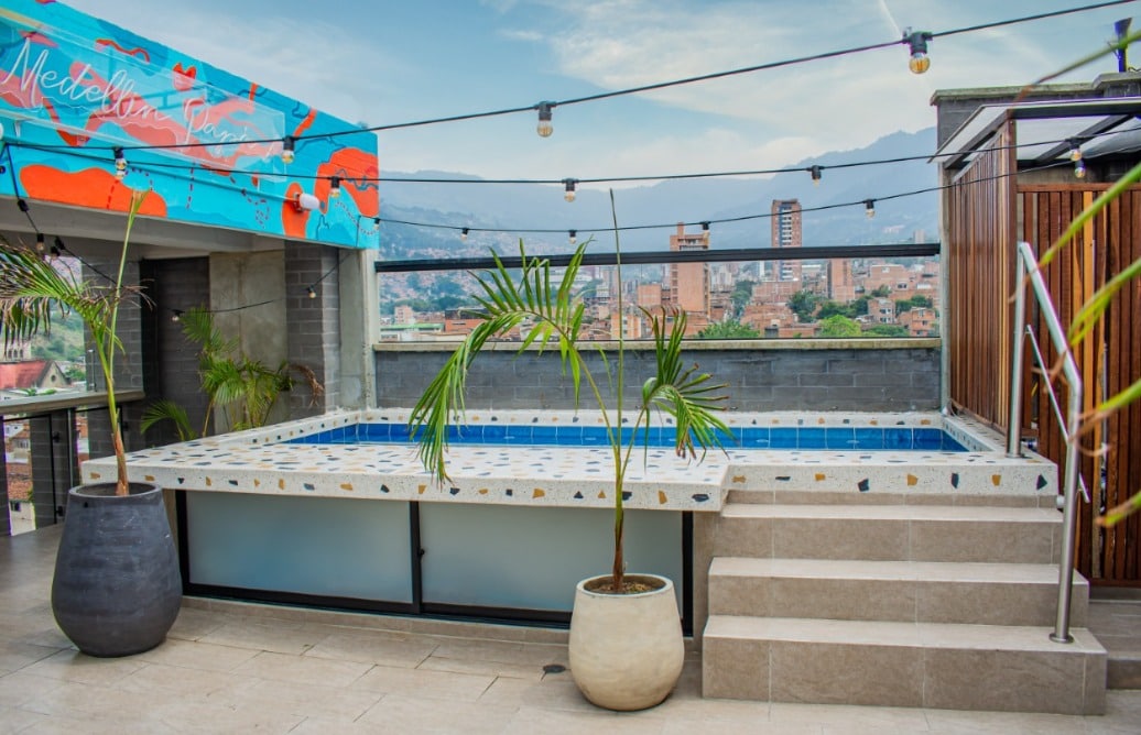 A rooftop area features a small pool surrounded by tile accents. Two large planters with greenery create a relaxed atmosphere. The backdrop includes a city view and a wall adorned with vibrant art, enhancing the outdoor experience.