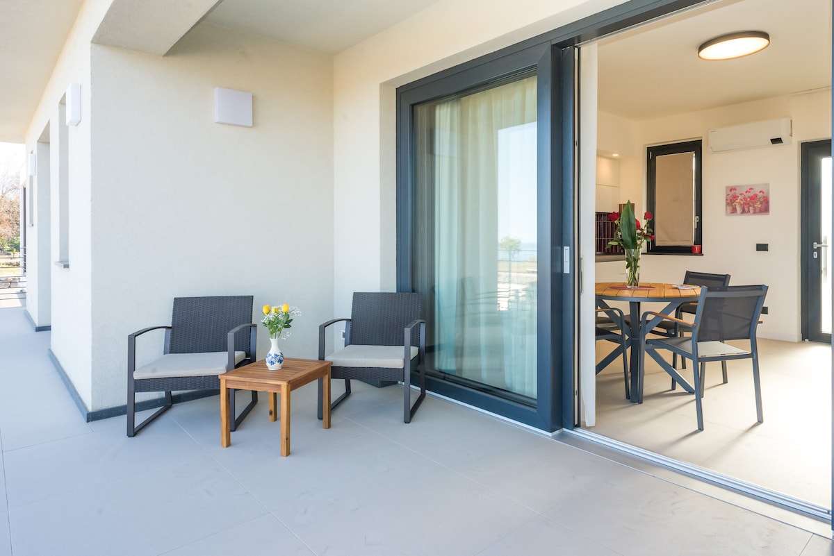A balcony area features two contemporary armchairs and a small wooden table with a vase of flowers. Large sliding glass doors provide a view into a well-lit interior space, where a dining table and chairs are visible, enhancing the connection between indoor and outdoor relaxation.