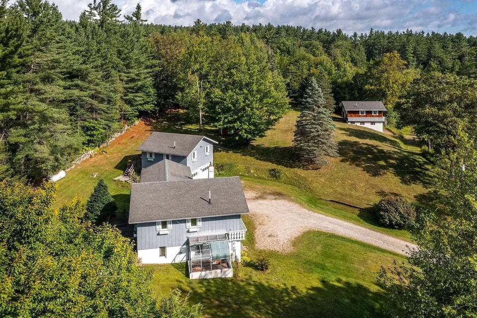 An aerial view showcases a serene property surrounded by greenery. The main building, with light grey siding and multiple windows, sits prominently on a grassy landscape. Nearby, a smaller structure is visible, framed by trees, with a gravel pathway leading through the area.