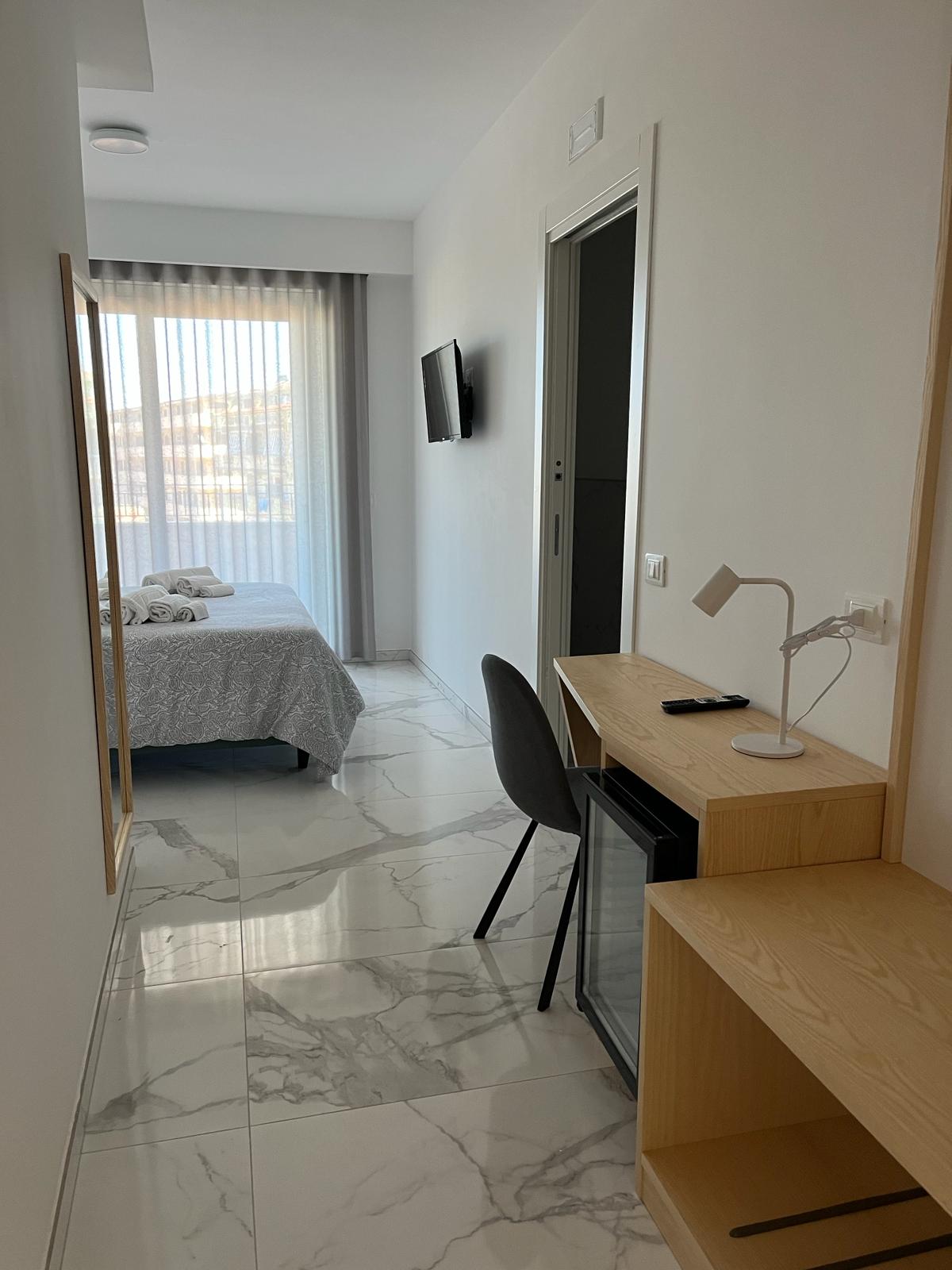 A well-lit corridor leads to a bedroom featuring a spacious bed with a light grey blanket. A small desk with a lamp and a chair is positioned next to a mini fridge. Natural light filters through sheer curtains covering a window at the end of the hallway.