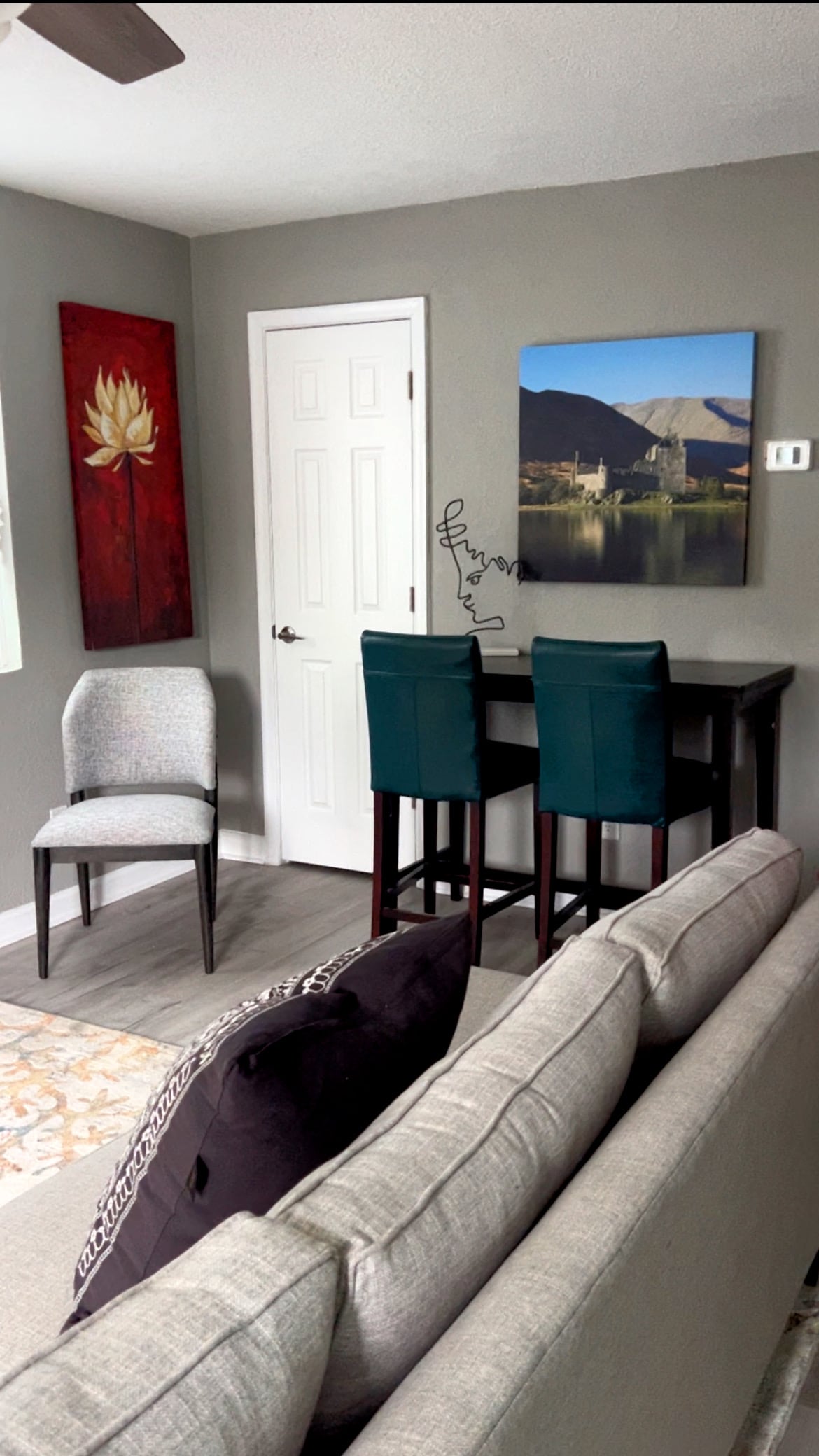 A cozy seating area features a light grey sofa and a small dining table with two teal chairs. A textured grey chair is positioned nearby. A large wall art piece displays a scenic view of mountains and water, while a vibrant flower painting adds color to the wall.
