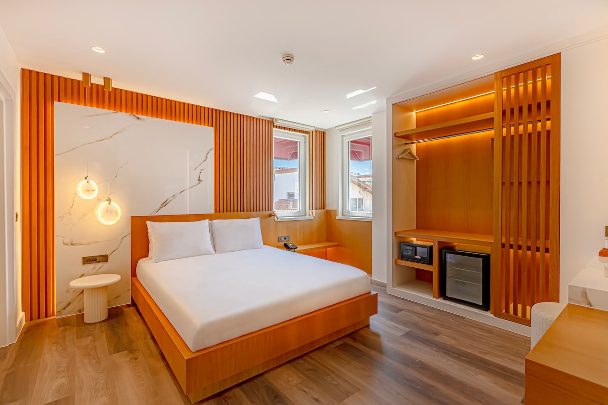 A modern guest room features a spacious bed with crisp white linens against a light wooden accent wall. Soft lighting is provided by two pendant fixtures, and a compact work area includes a microwave and refrigerator. Natural light enters through large windows with minimal coverings.