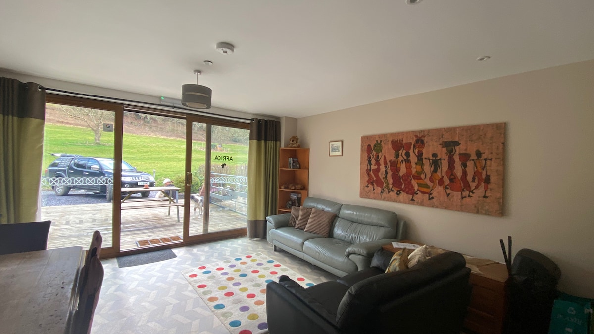A living area is revealed with modern furnishings and a colorful area rug. Large glass doors lead to an outdoor space, allowing natural light to fill the room. A wall adorned with a vibrant art piece adds cultural character, while seating arrangements invite relaxation.