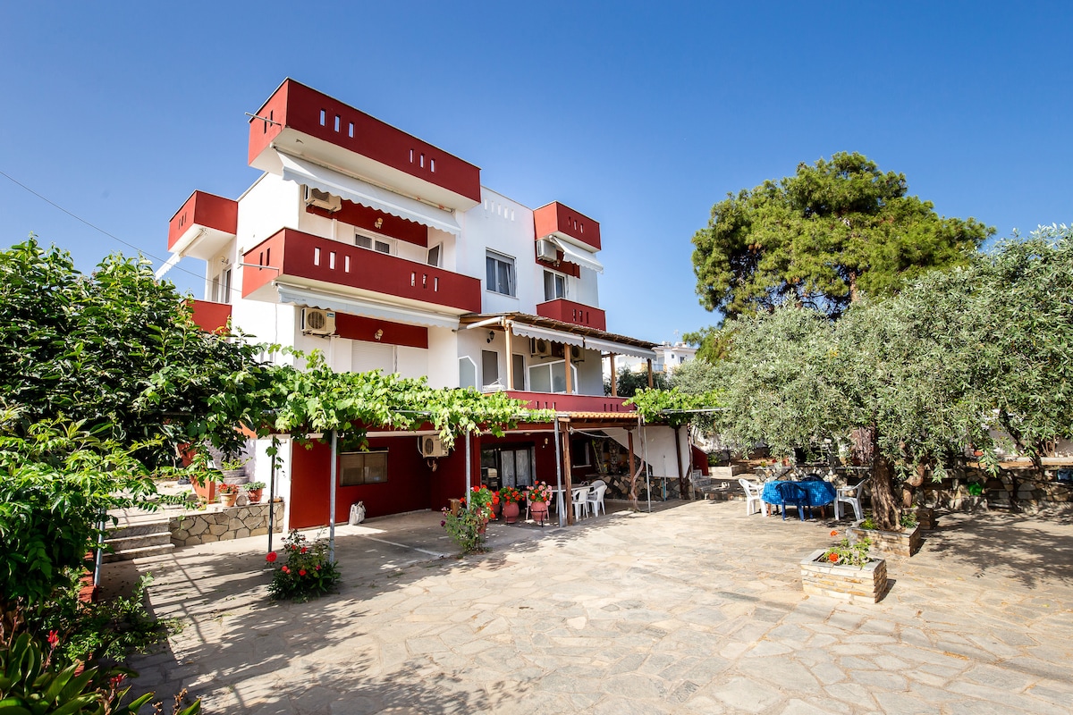 The exterior of the family-run property showcases a three-story building with red and white façades. Surrounding greenery includes lush trees and flowering plants, creating a pleasant outdoor environment. A spacious stone-paved area is visible, accommodating outdoor furniture and parking.