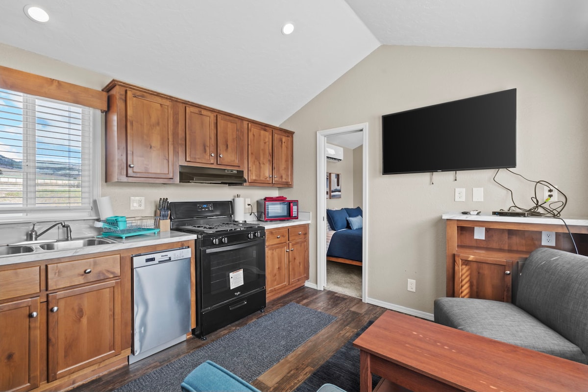 A compact kitchen area features wooden cabinetry and modern appliances, including a stove and microwave. A dining table is positioned near a window with natural light illuminating the space. A seating area with a gray couch is arranged near a television mounted on the wall.
