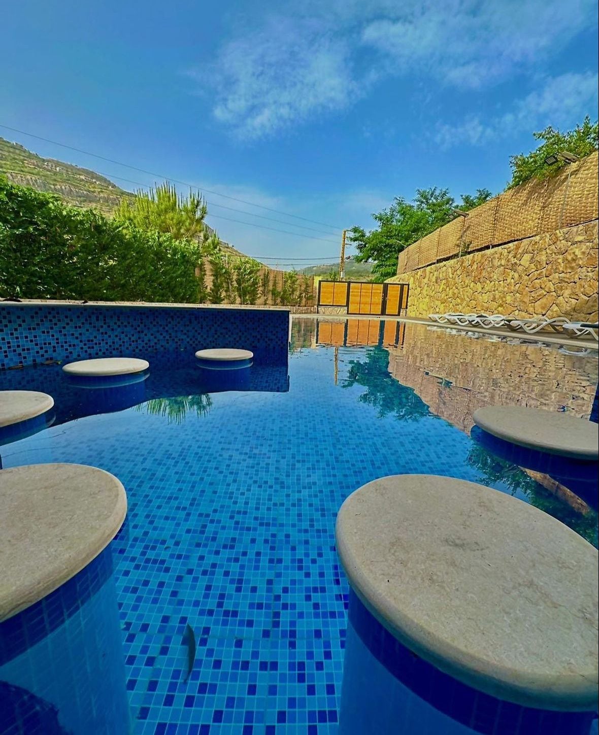 A tranquil pool area features circular stone steps leading into the water, surrounded by vibrant blue tiles. The clear water reflects the sky and surrounding greenery, creating a serene environment. Lush trees and a rocky wall frame the space, enhancing the natural setting.