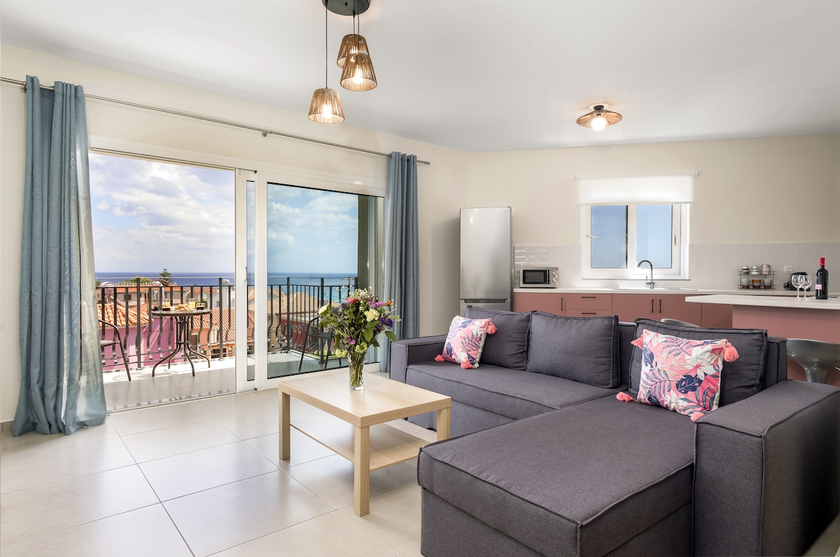 A modern living area features a spacious gray sofa adorned with floral accent pillows. Natural light floods the space through sliding glass doors, which open to a balcony with sea views. A stylish coffee table sits in front of the sofa, and a sleek kitchen can be seen in the background.