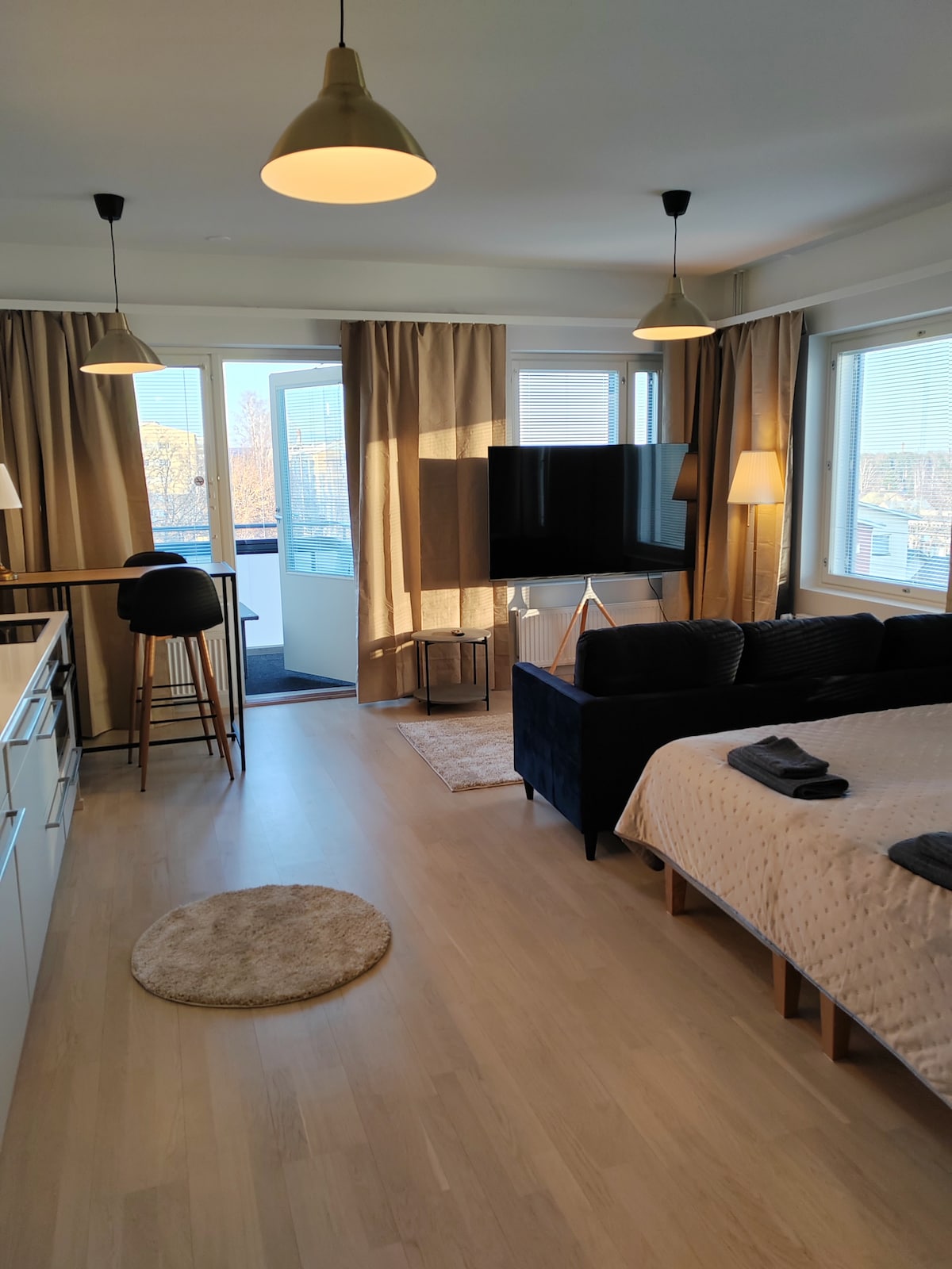 The studio features a cozy seating area with a dark sofa and a large TV. Soft lighting is provided by pendant lamps and natural light enters through floor-to-ceiling windows. A small balcony is visible in the background, complementing the inviting atmosphere of the space.