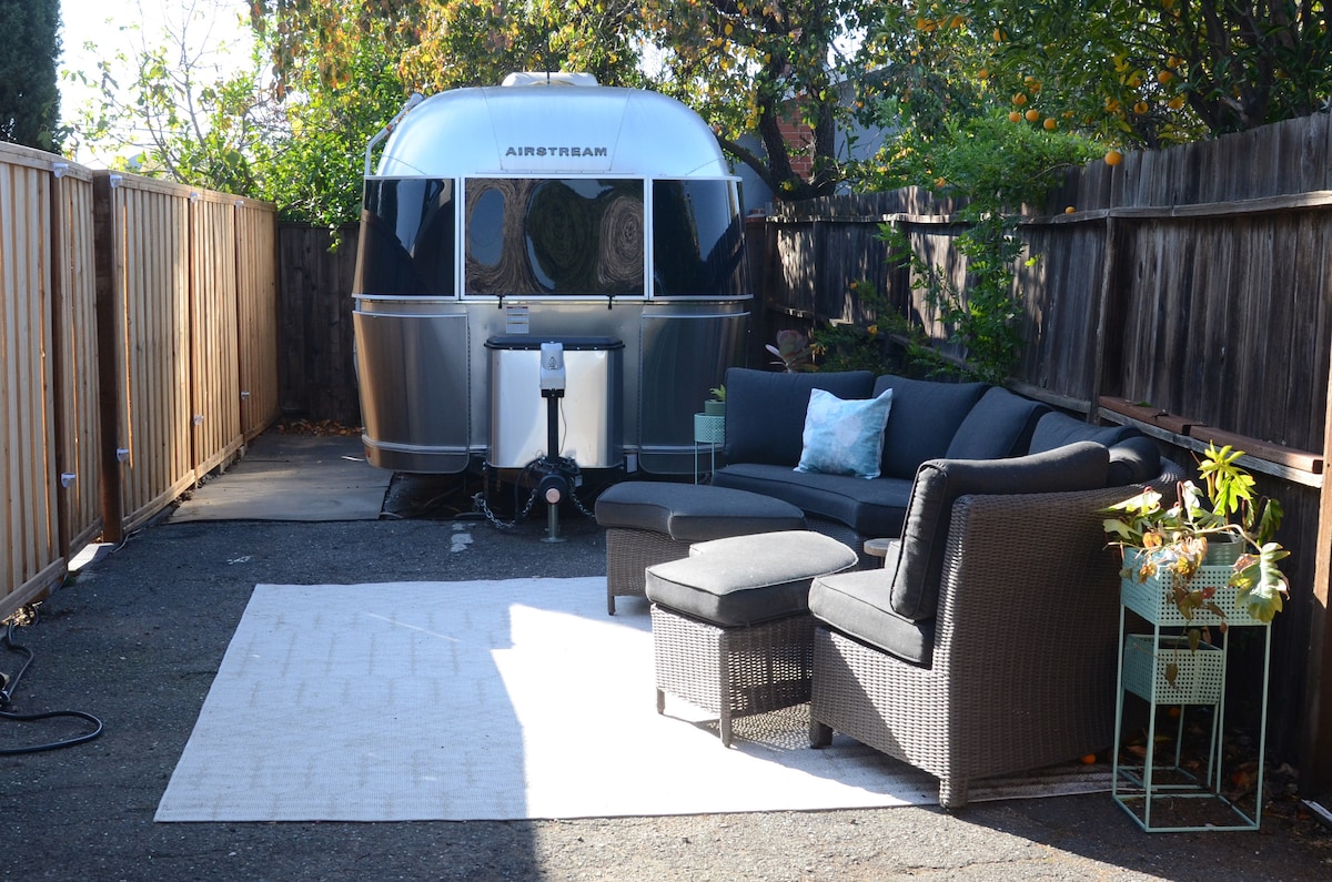 An outdoor seating area features a dark sectional couch and a matching lounge chair, placed on a light area rug. A small metal table holds plants beside the Airstream, which sits enclosed by wooden fencing and surrounded by greenery.