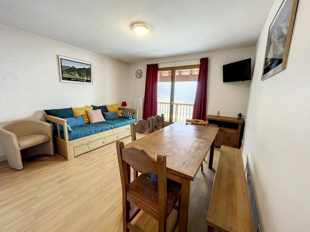 A spacious living area features a convertible couch adorned with colorful cushions and a wooden dining table surrounded by chairs. Large doors open to a balcony, inviting natural light and showcasing scenic views of the outdoors. A flat-screen TV is mounted on the wall.
