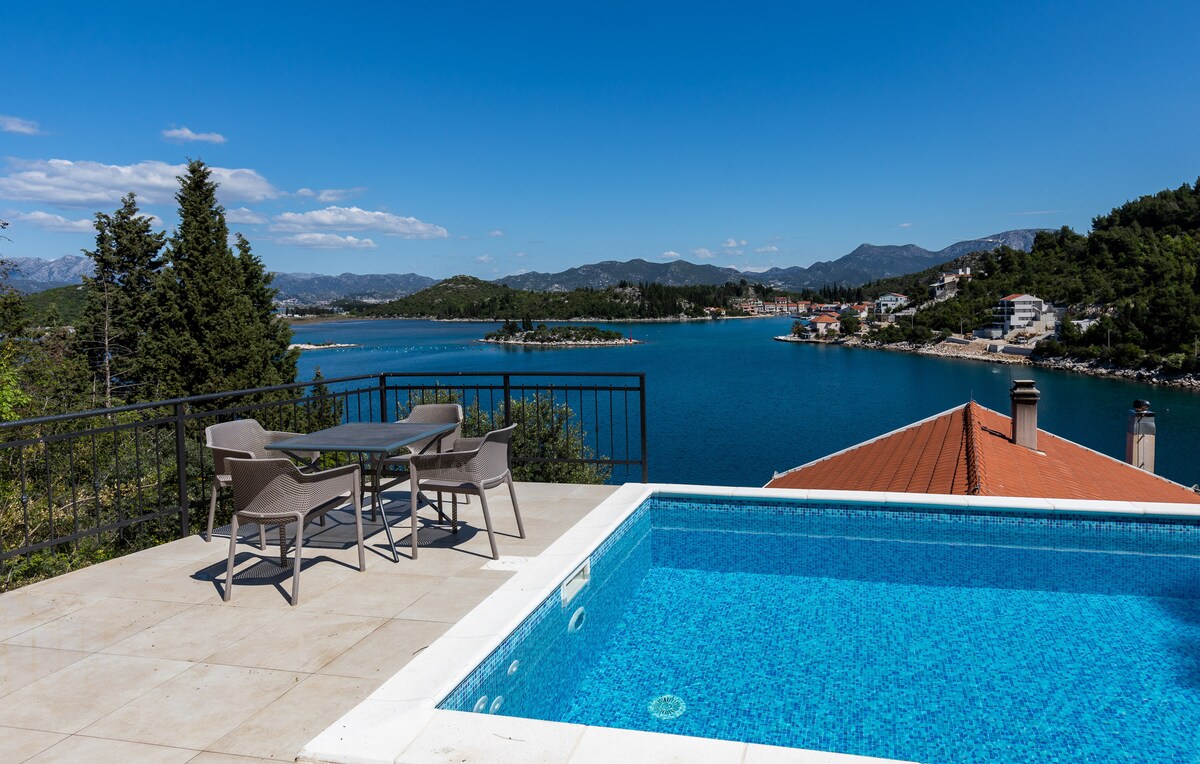 An outdoor terrace features a private swimming pool and a dining set composed of four chairs surrounding a table. The scene is complemented by panoramic views of the turquoise water and hills, creating a serene environment for relaxation.