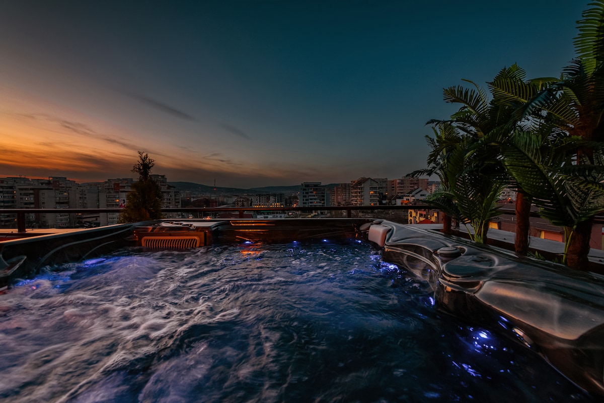 A jacuzzi offers a relaxing setting with swirling water illuminated in a soft blue glow. The terrace features palm plants and showcases a panoramic view of the city against a twilight sky, enhancing the tranquil atmosphere.