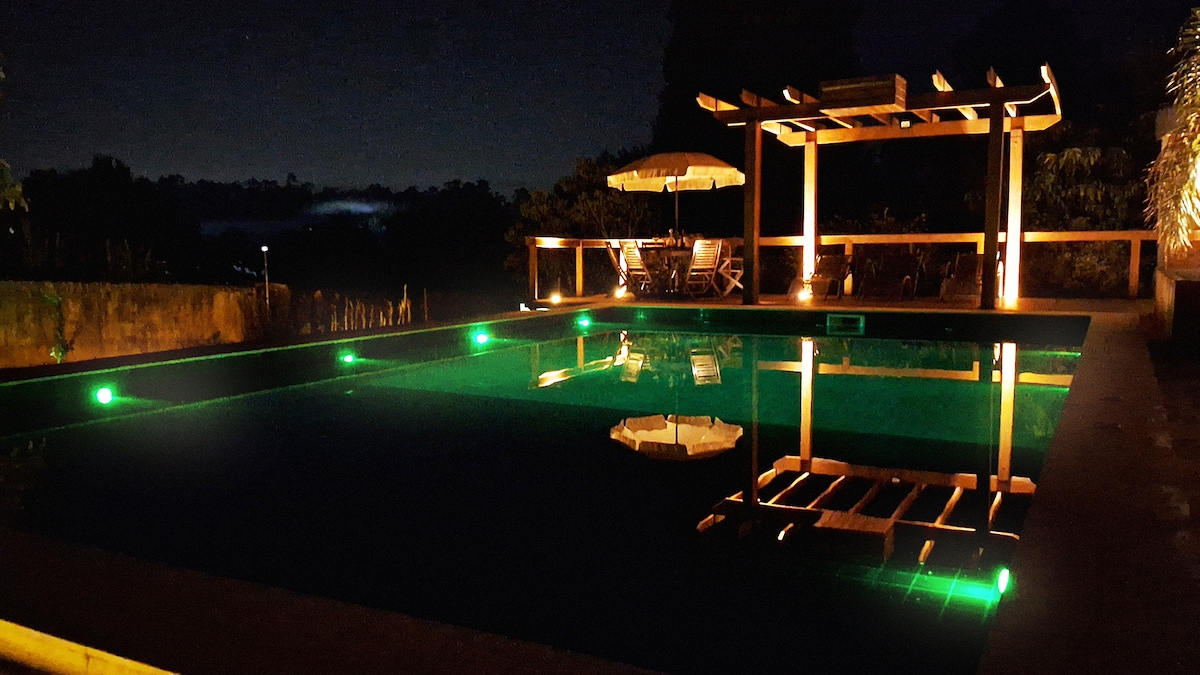 A tranquil pool area is illuminated by soft, green lights against a dark backdrop. Wooden deck seating can be seen, enhancing the outdoor space's inviting atmosphere. The water reflects the gentle lighting, creating a serene ambiance at night.