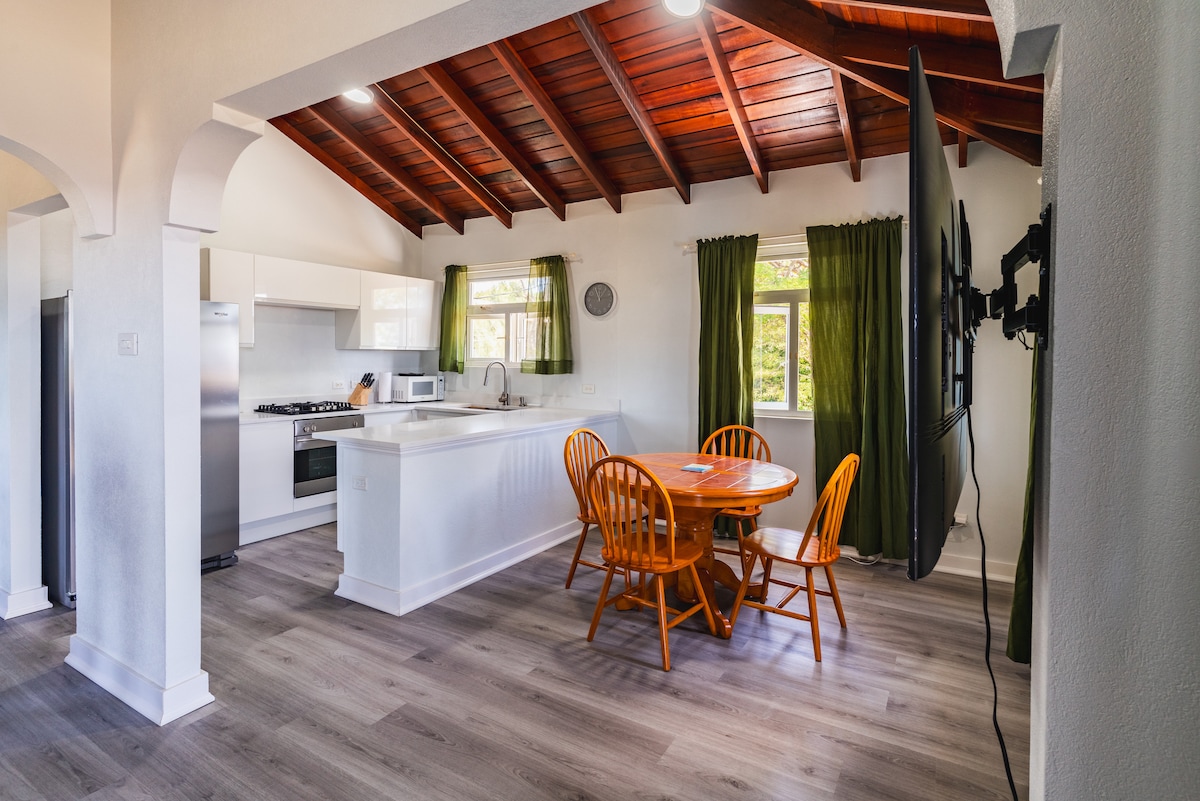 The open-plan kitchen and dining area feature a wooden ceiling with exposed beams. A round wooden dining table with four chairs is set beside a window, allowing natural light to illuminate the space. The kitchen is equipped with modern appliances, including a full fridge and stove.