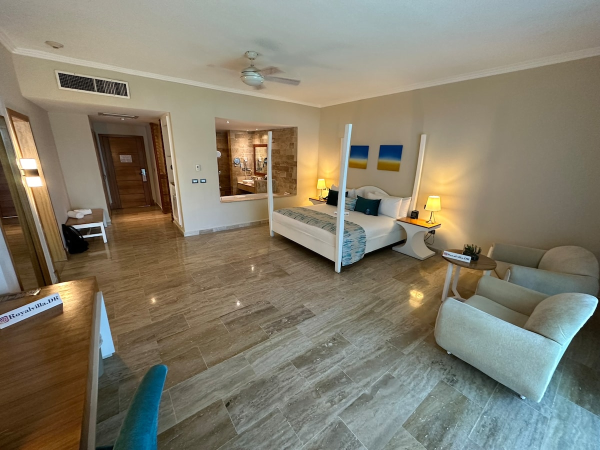 A spacious room features a king bed with a light-colored headboard, complemented by two bedside lamps. A large mirror reflects the cozy seating area, which includes two light armchairs and a desk. The room is illuminated by natural light from a nearby window.