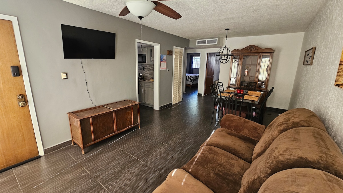 A spacious living area features a large, comfortable sofa and a wall-mounted television. A dining table with eight chairs is situated nearby, and an entryway leads to the kitchen and bedrooms. Warm light complements the neutral-toned walls and tiled flooring.