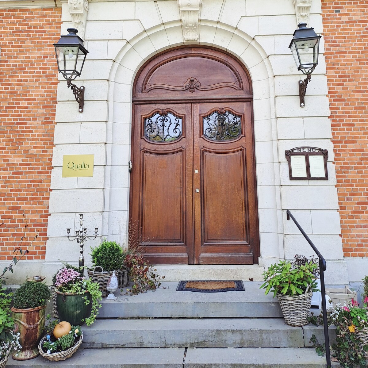 A grand wooden door with intricate carvings stands as the entrance to the building, flanked by decorative wall lanterns. A series of potted plants and a candelabra are positioned along the steps, adding to the inviting approach to the entrance.