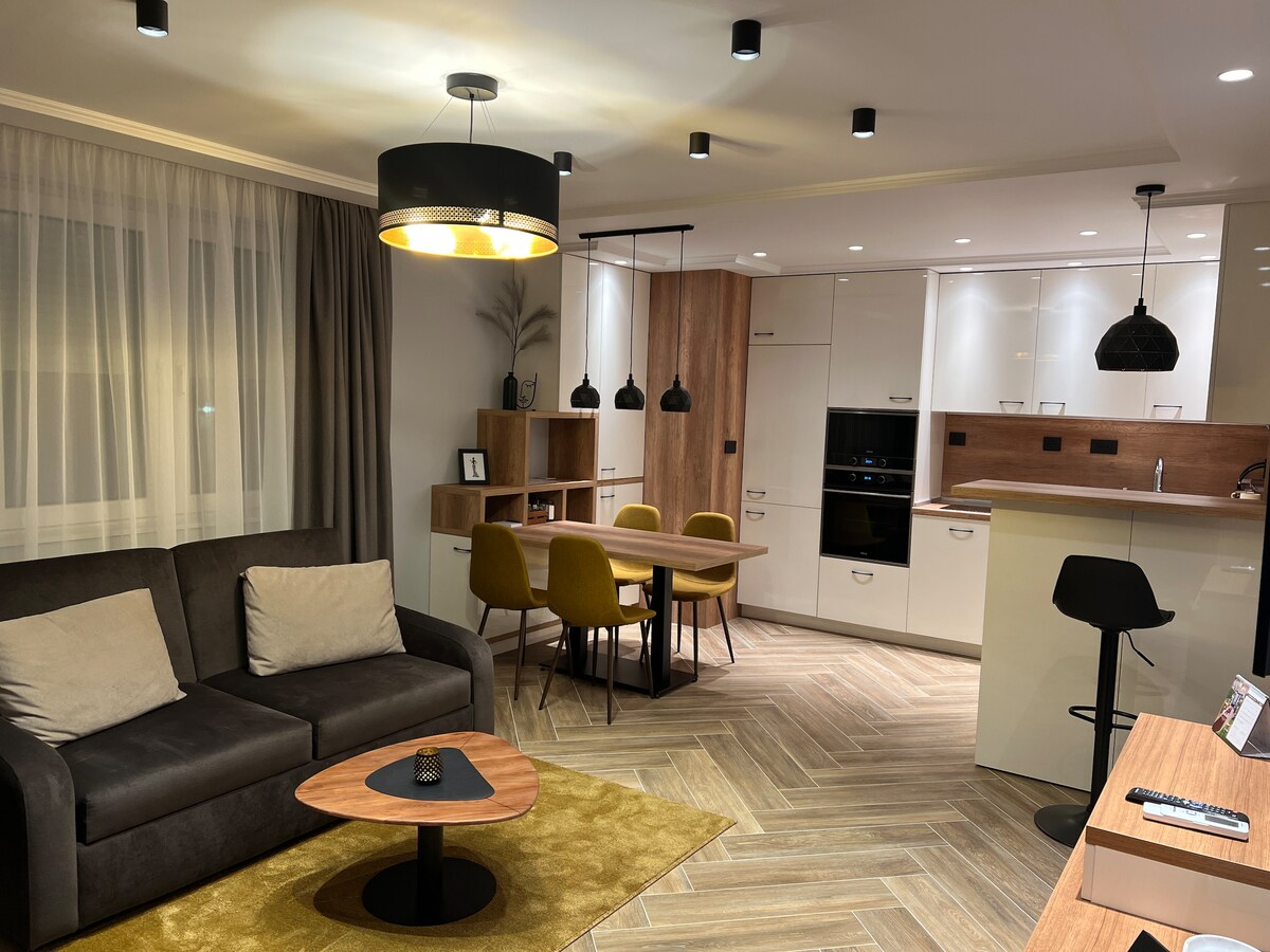 A modern living area features a comfortable dark sofa with light-colored cushions, complemented by a wooden coffee table. A dining area with four yellow chairs and a stylish pendant light is visible. The kitchen showcases sleek cabinetry and contemporary appliances under soft lighting.
