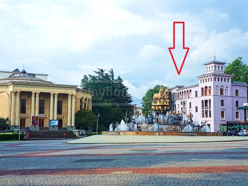 A city square features a prominent fountain adorned with sculptures, surrounded by lush greenery. Two architecturally distinct buildings are visible: a grand theater with impressive columns on the left and an elegant pink structure on the right. The scene reflects a vibrant urban setting.