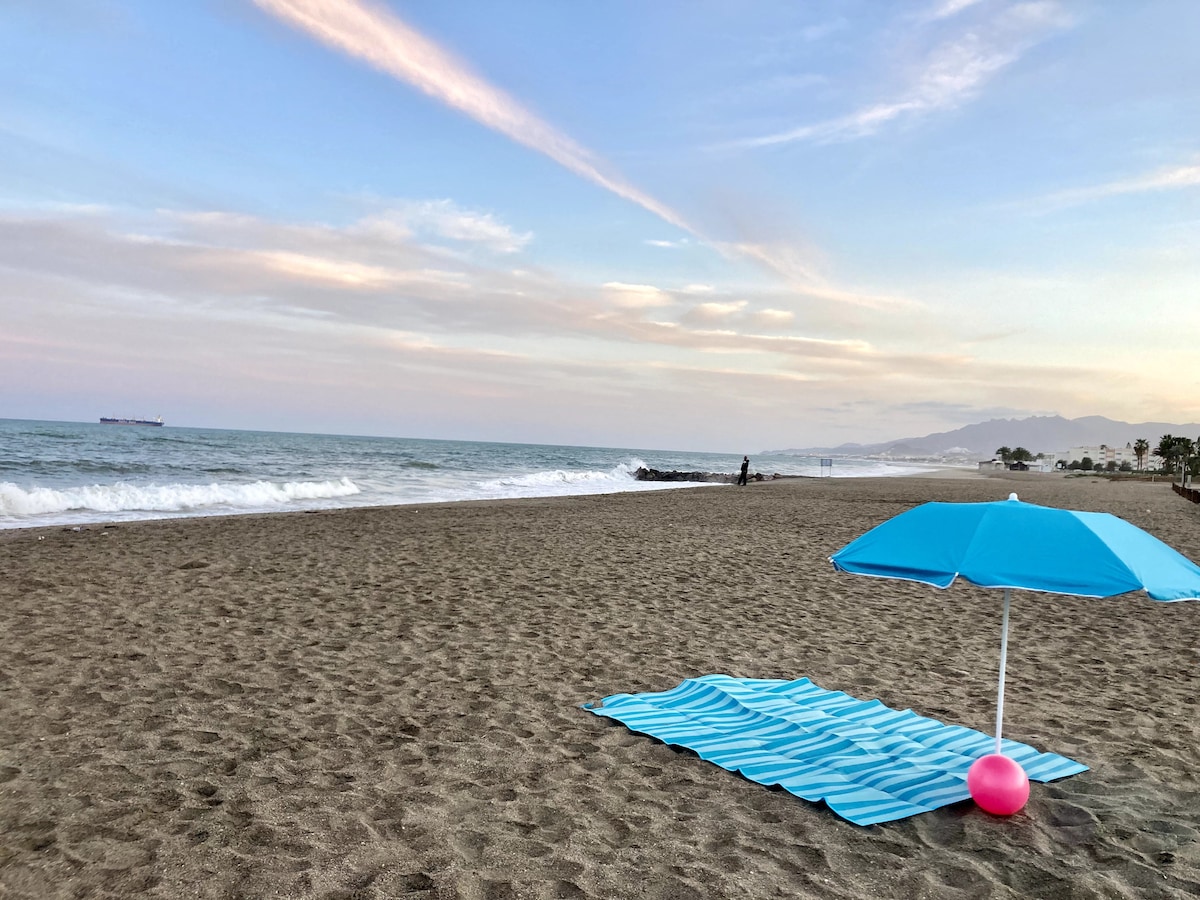A sandy beach is visible, featuring a blue mat laid out on the shore. A bright blue umbrella stands nearby, providing shade over the mat. Gentle waves can be seen lapping at the beach, with distant mountains on the horizon and a ship in the water.
