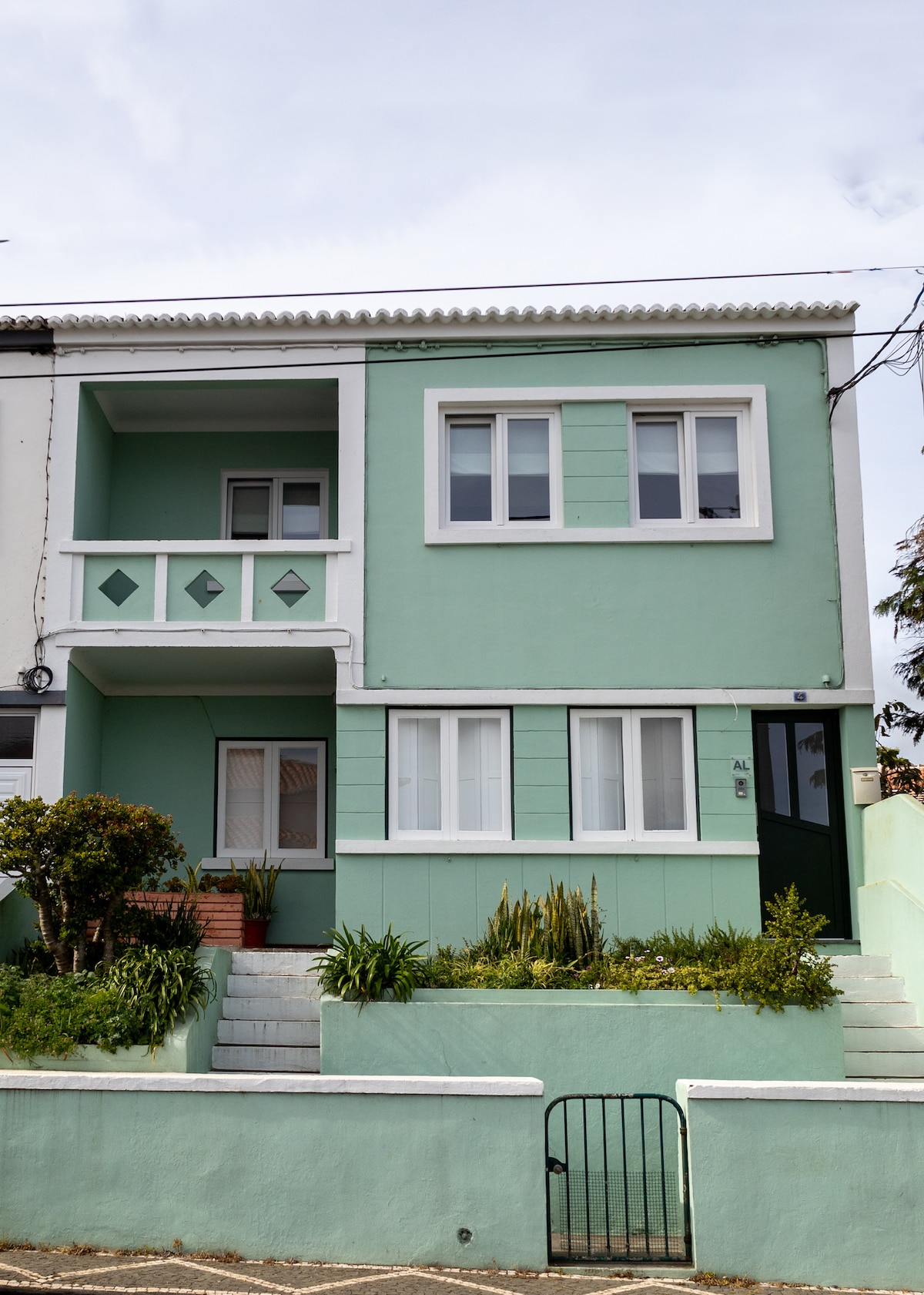 A two-story building features a soft green exterior with balcony accents and simple geometric designs. Surrounding the entrance, a low wall encloses a small garden with various plants, and steps lead up to the front door. Light clouds can be seen in the sky above.