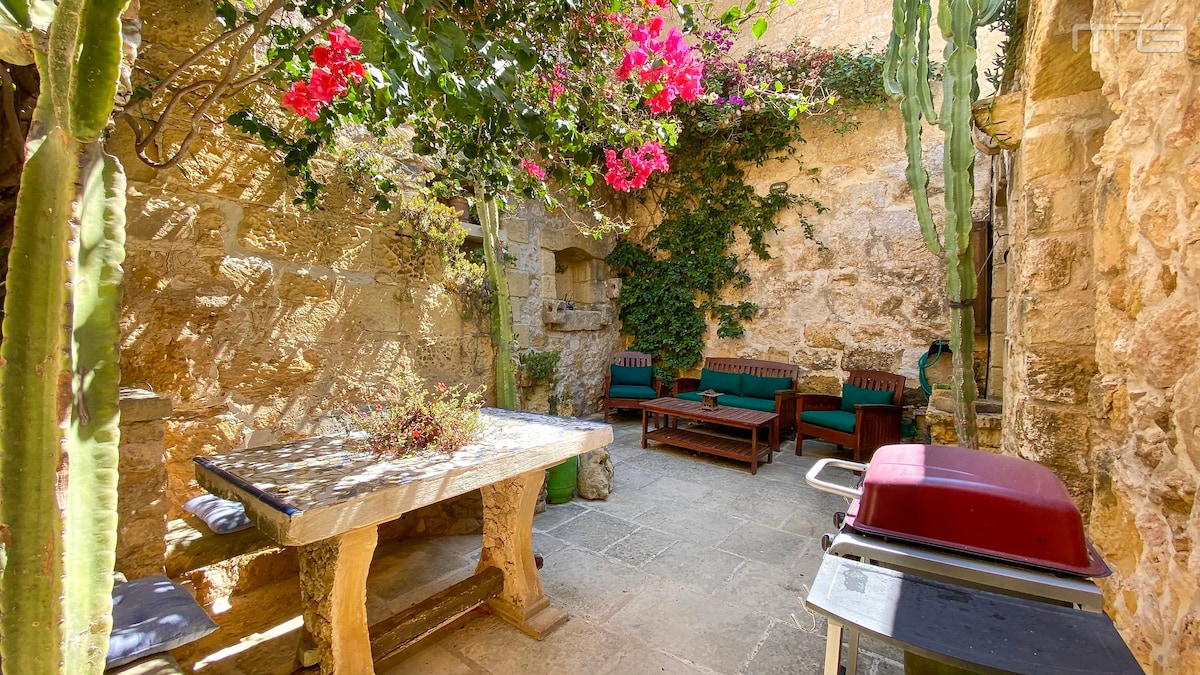 A secluded courtyard area is featured, surrounded by stone walls and vibrant greenery. Comfortable seating is arranged on wooden benches, with a rustic stone table positioned nearby. A barbecue grill is visible, along with blooming flowers adding color to the tranquil setting.