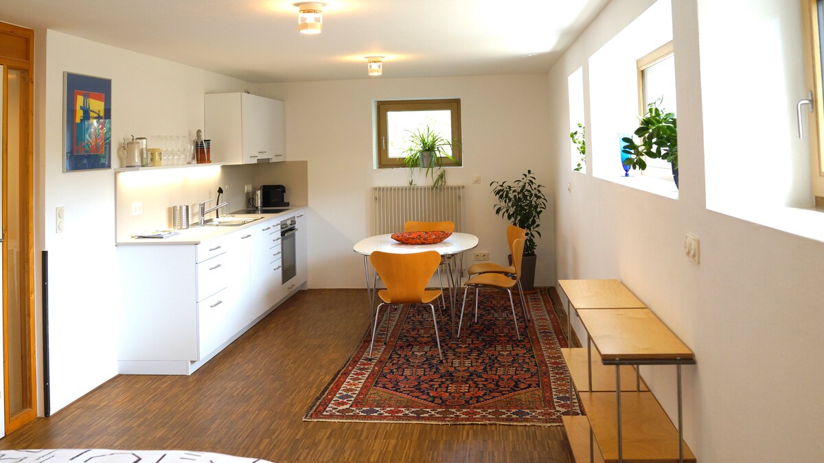 A bright, modern kitchen area features sleek white cabinets and appliances. A round dining table with four chairs is positioned on a patterned rug. Natural light enters through two small windows, and a potted plant adds a touch of greenery to the space.