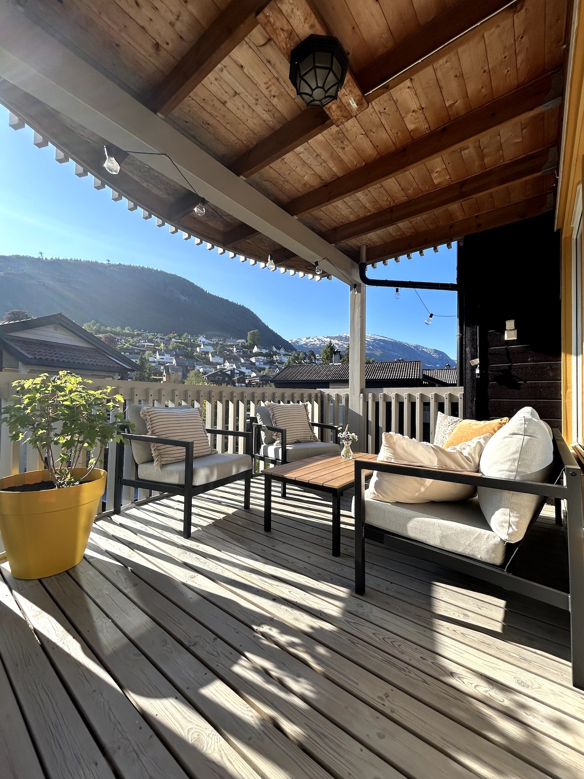 A covered balcony is presented with wooden flooring and a sloped ceiling. Two chairs and a small table provide seating options. A potted plant adds a touch of greenery, while views of mountains are framed in the background, illuminated by natural light.