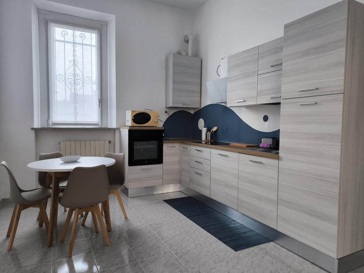 A modern kitchen features light wood cabinetry and a sleek design. An oven is integrated into the cabinetry, while a round dining table with four chairs is positioned nearby. Natural light enters through a window, highlighting the minimalist decor and blue accent wall.