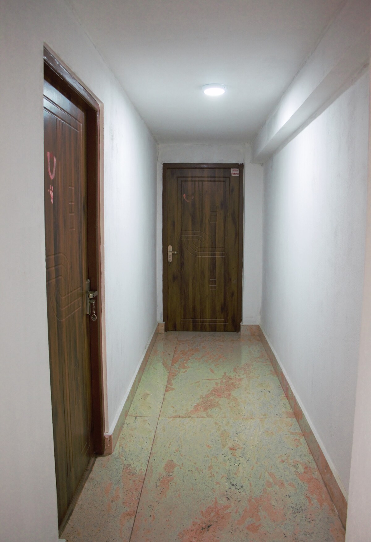 A well-lit corridor features smooth, polished flooring with a blend of colors. Two wooden doors are positioned on opposite sides, leading to different accommodation units. White walls enhance the sense of space, creating a clean and welcoming environment.