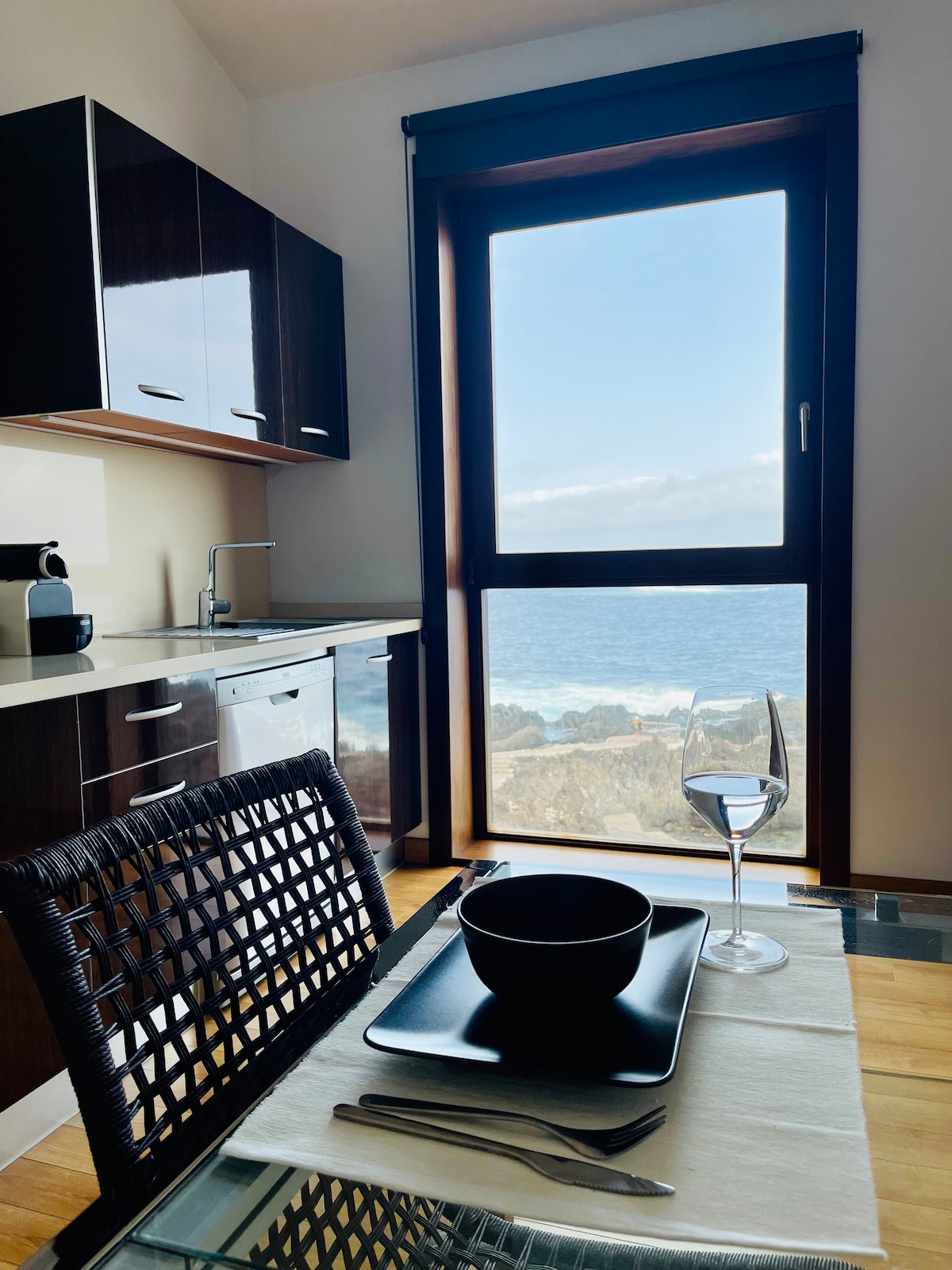 A kitchen area features modern cabinetry and appliances, complemented by a dining table set for two. A large window provides a view of the ocean, allowing natural light to illuminate the space. A black bowl and glass are placed on the table, ready for dining.