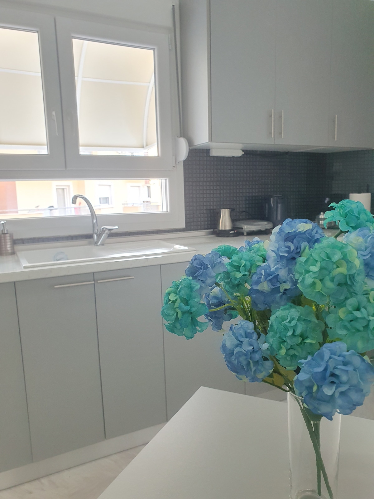 A modern kitchen is highlighted by sleek grey cabinetry and a white countertop. A sink with an overhead window allows natural light to fill the space. A vibrant blue flower arrangement sits on the table, adding a touch of color to the minimalist decor.