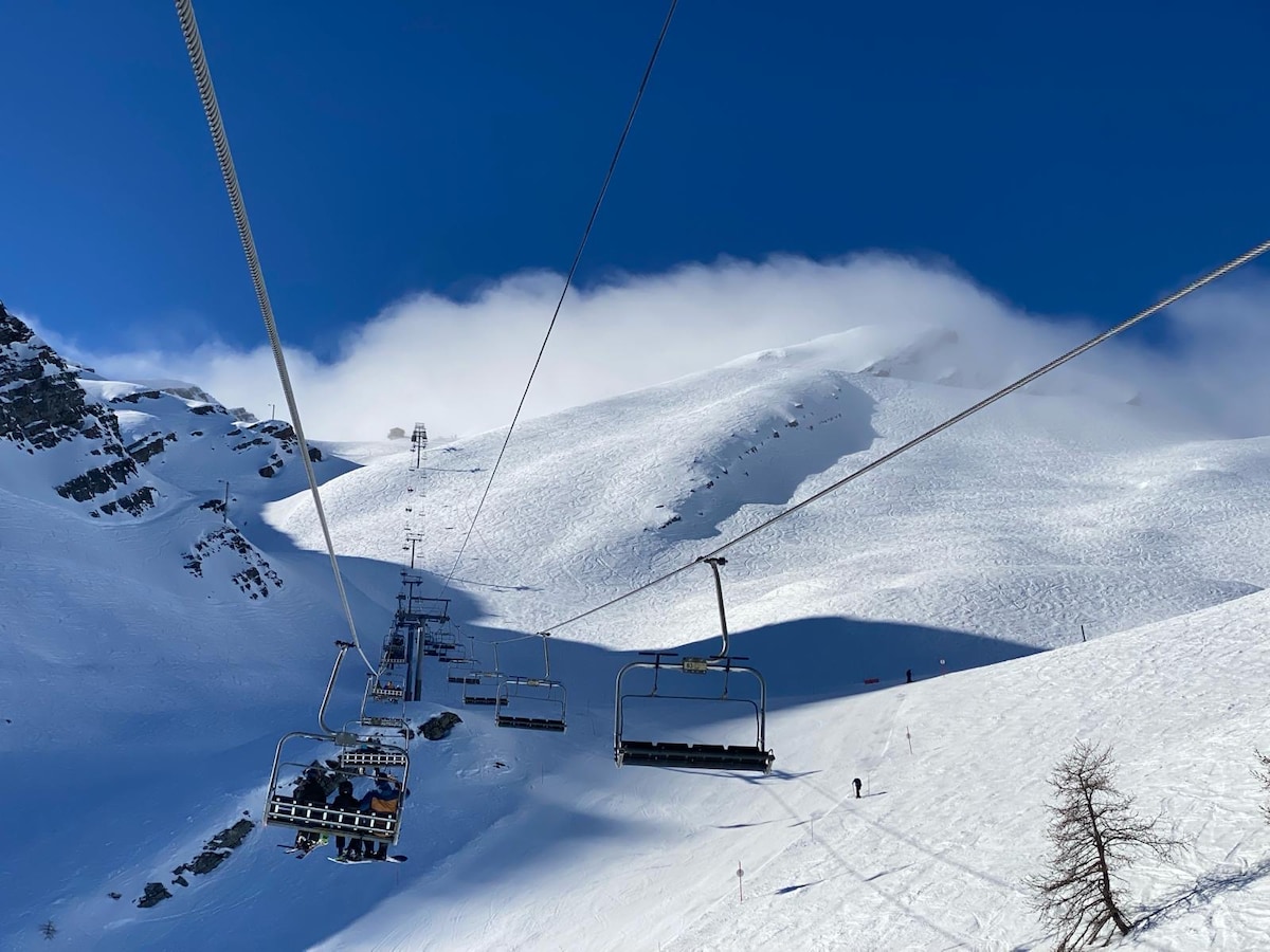 A ski lift ascends over a snow-covered landscape, with slopes blanketed in fresh powder. The backdrop features a mountainous terrain under a clear blue sky, while a few skiers can be spotted on the trails below.