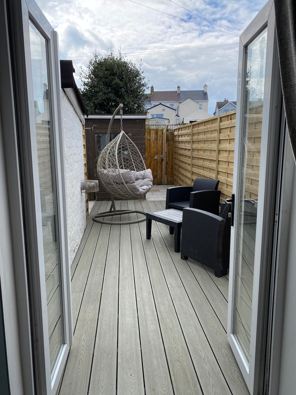 A private outdoor space is highlighted by a swinging chair and two black lounge chairs, arranged on a wooden deck. The area is enclosed by wooden fencing for added privacy, with plants visible in the background, creating a calm retreat.