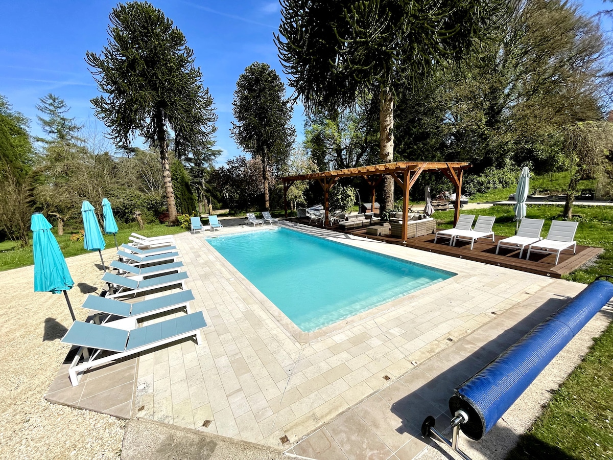 A spacious outdoor pool is framed by lounge chairs and umbrellas, ideal for sunbathing. A wooden pergola offers shade beside the pool, surrounded by lush greenery and mature trees, creating a relaxing environment for guests.