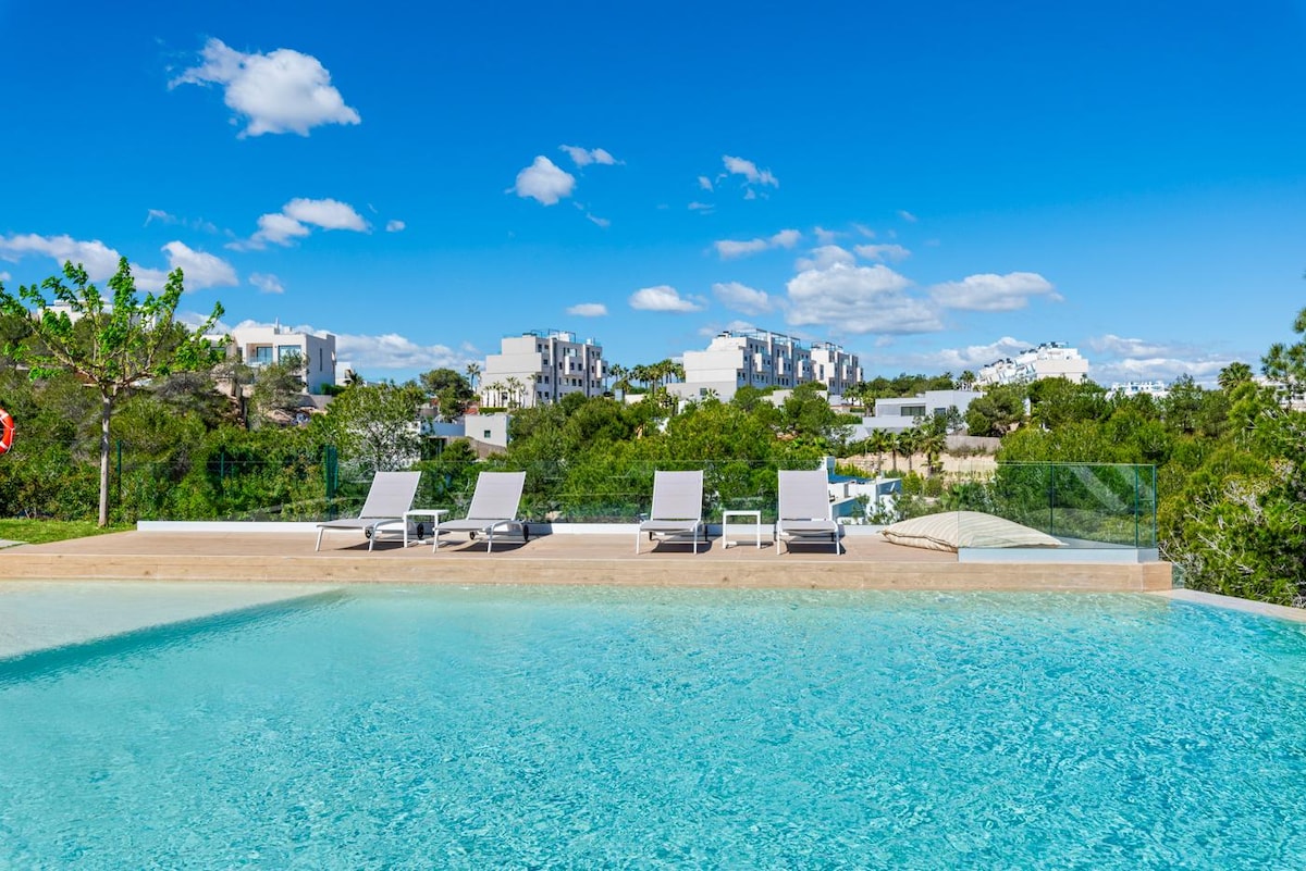 An inviting outdoor pool reflects the clear blue sky, surrounded by a spacious deck fitted with several lounge chairs. Lush greenery and distant buildings create a tranquil backdrop, enhancing a sense of privacy and relaxation.
