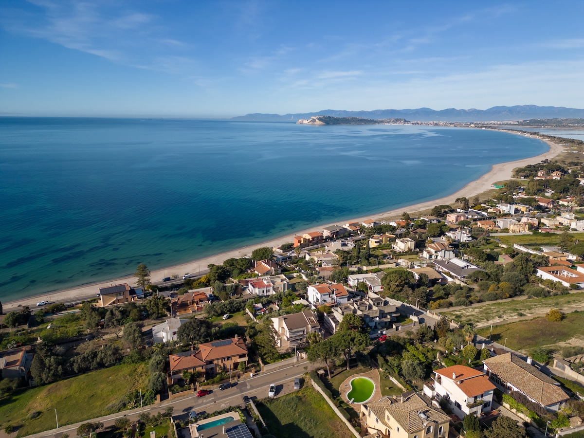 Aerial view of a coastal area featuring a serene beach and crystal-clear waters. The shoreline is lined with residential homes, surrounded by greenery. In the distance, mountains are visible under a blue sky, enhancing the scenic coastal landscape.