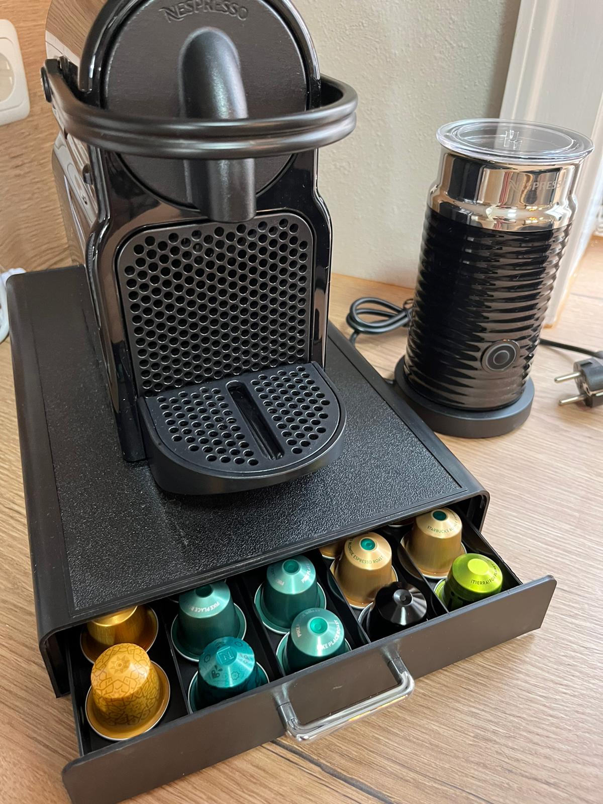 A Nespresso coffee machine is positioned on a wooden surface, accompanied by a milk frother and a drawer containing various colorful coffee capsules. The setup is organized, showcasing a modern and functional design for coffee preparation.