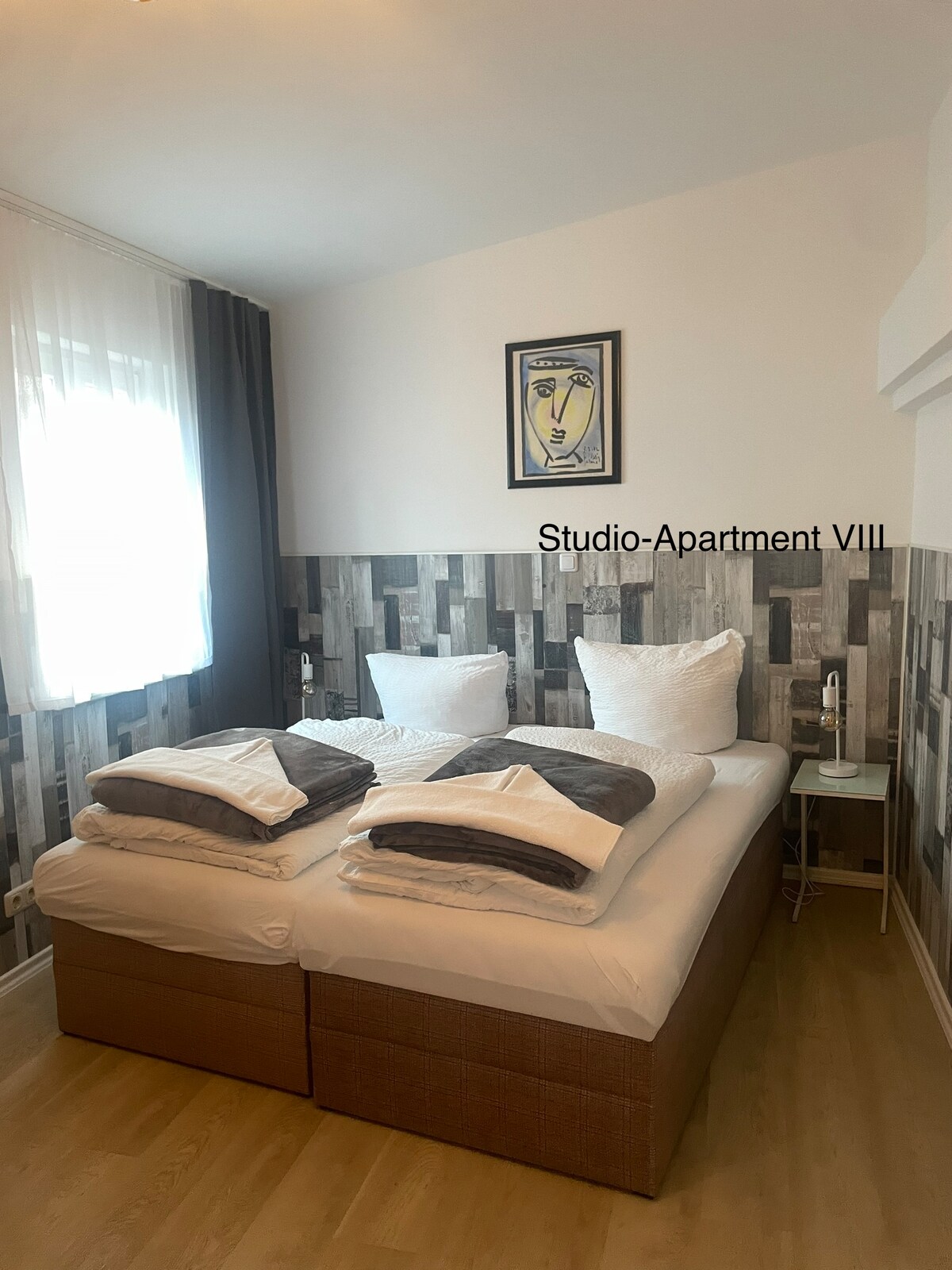 The sleeping area features a comfortable bed arranged with multiple pillows and beddings. Light enters through sheer curtains in front of a window. A decorative art piece is mounted on the wall above the bed, adding a touch of character to the room.